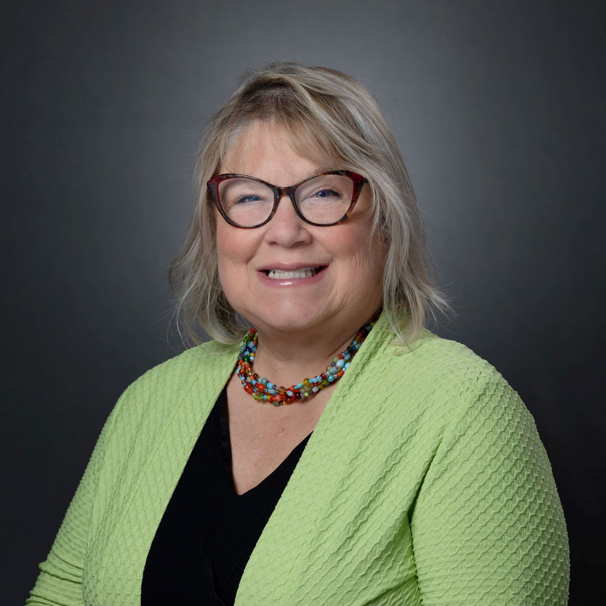 Headshot of Sherrie Smith female with shoulder length blonde hair and glasses wearing green sweater