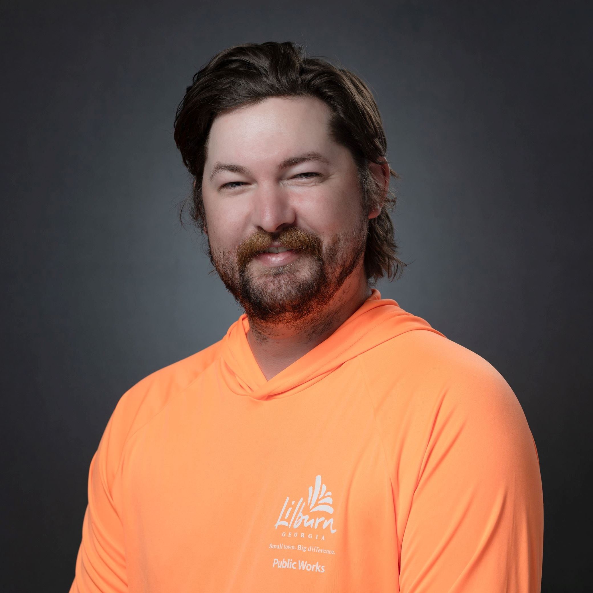Headshot of Colin Conroy male with longer brown hair and beard/mustached wearing orange tee